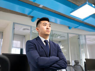 Serious young Asian male manager in formal suit looking away while standing near table with computers in contemporary coworking office