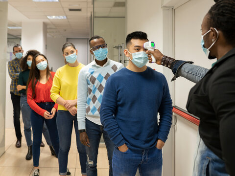 Group of multiracial colleagues in casual clothes and medical masks passing through procedure of body temperature control for coronavirus prevention before entering workplace