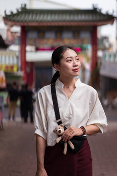 Positive young Asian female traveler in casual clothes with cute little toy in hand standing on urban square and looking away while exploring streets of Tainan city in Taiwan
