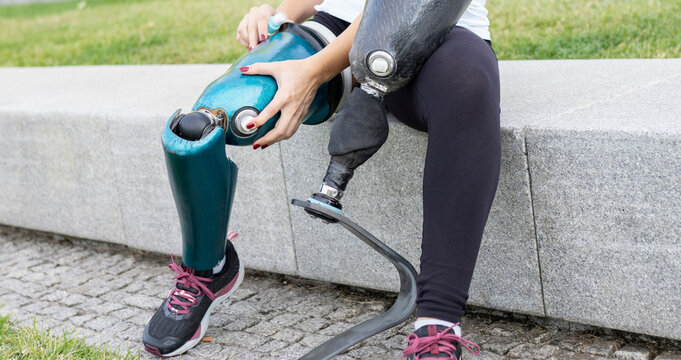 Unrecognizable crop female runner changing leg prosthesis for training while sitting on stone border in urban park