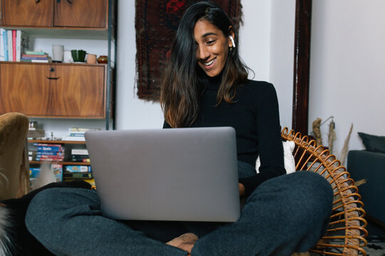 Cheerful Indian female sitting in wicker chair and having video call on laptop while smiling
