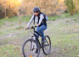 Full body of positive young female in activewear and helmet riding bicycle through autumn forest with golden foliage in sunny day in countryside