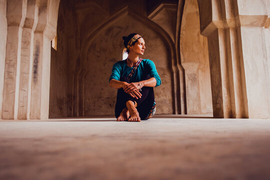 Full body of peaceful young barefooted ethnic female traveler in traditional clothes sitting on floor in ancient oriental palace in India and looking away thoughtfully