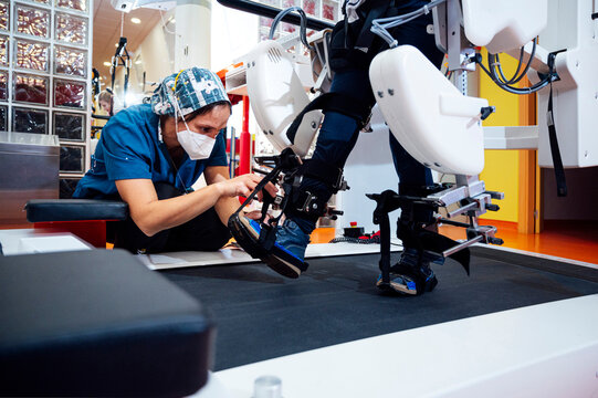 Female specialist in protective mask fastening belts on legs of unrecognizable patient with disability while preparing for physiotherapy exercise on modern equipment in rehabilitation center