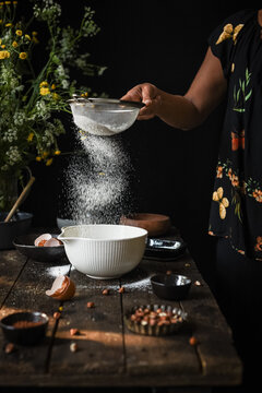 Crop anonymous female sieving flour into bowl while preparing dough for pastry at wooden table