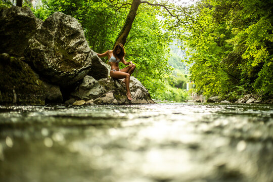 Side view of alluring young fit female traveler in bikini sitting on rocky cliff near lake in green lush rainforest in Fuente De