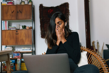 Low angle of astonished Indian female sitting with laptop and reading news online while covering mouth