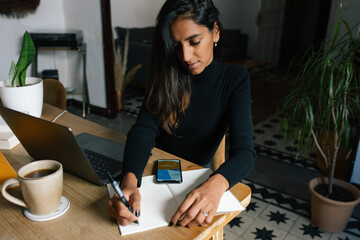 High angle of Indian female entrepreneur sitting at table and writing plans in notebook while working at home in cozy workplace