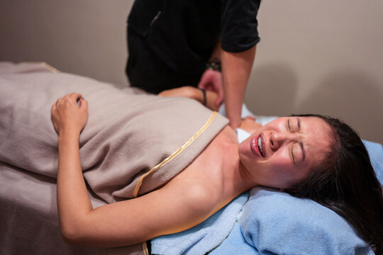Side view of young Asian female patient crying in pain while getting professional osteopathic massage from therapist in clinic