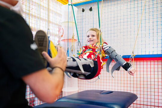 Crop anonymous specialist giving support to girl with Angelman syndrome during rehabilitation training with elastic straps
