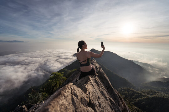 Back view of female ethnic hiker sitting on top of Yuanzui Mountain and taking self portrait on smartphone