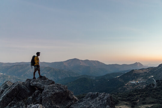 Full body side view of lonely unrecognizable male explorer with backpack standing on top of rough rocky mountain and enjoying freedom while hiking through highlands at sunset