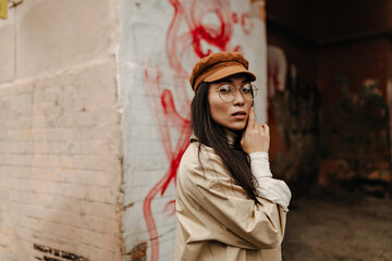 Asian girl in cap and coat posing near abandoned house