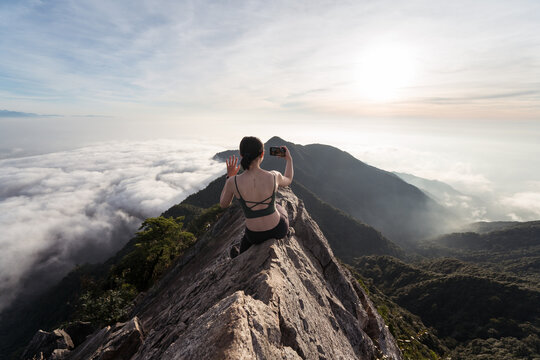 Back view of female ethnic hiker sitting on top of Yuanzui Mountain and taking self portrait on smartphone