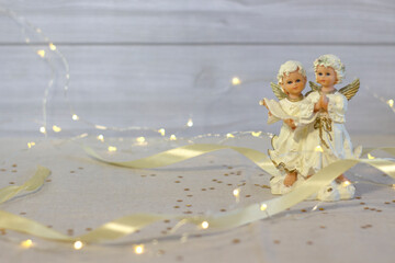 Little ceramic angels pray and sing songs, on a blurred background, satin ribbon and lights