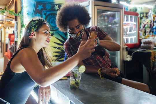 African American man showing photo on mobile phone to girlfriend while standing near counter of outdoor beach bar - Powered by Adobe