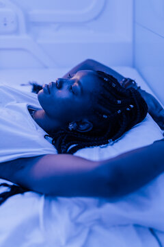 Young black woman resting on bed in contemporary capsule hotel with futuristic interior design and blue illumination looking up