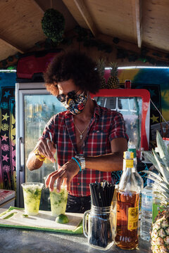 African American barman in sunglasses and protective mask mixing alcoholic drink with lime and ice while preparing order for client in outdoor bar