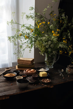 Set of various ingredients for sweet chocolate pastry recipe arranged on wooden table with fresh flowers in vase near window