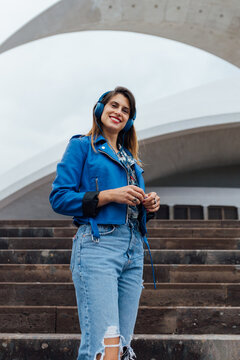 Low angle of happy young female in trendy jacket and ripped jeans looking at camera while standing on stairs near urban building and listening to music through headphones