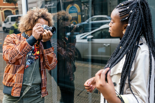 Creative male photographer with camera shooting photo of stylish young African American female model with afro pigtails near glass wall of urban building