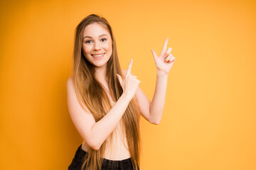 A cute caucasian girl with long hair in a beige top and black jeans smiles against an orange background and points her fingers to the right. space for text