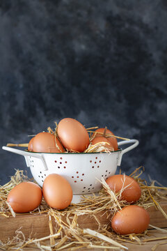 Composition with brown chicken eggs in white bowl placed on wooden board with dry hay against dark background