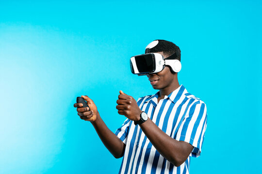 Side view of young African American male in VR goggles and with joystick experiencing virtual reality on blue background in modern studio