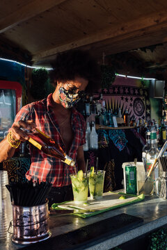 Black male bartender in protective mask adding alcohol into glass with ice and lime while preparing tropical cocktail at counter of beach bar