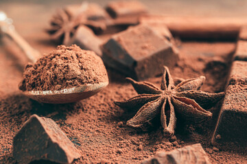 Brown cocoa powder in the spoon, chopped chocolate cubes and star anise on dark background