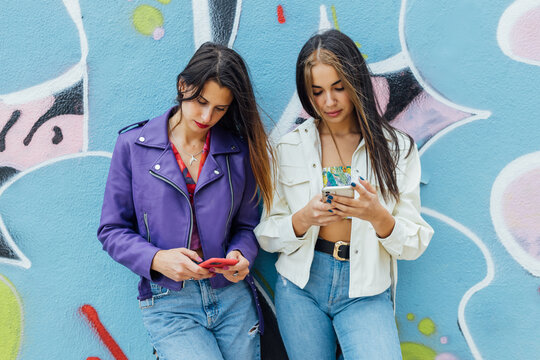 Anti-social trendy millennial women in stylish wear standing near colorful painted wall browsing on smartphone