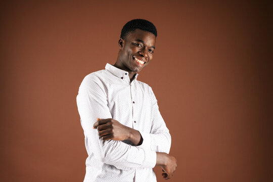 Delighted handsome African American male wearing classic white shirt standing on brown background in studio and looking at camera