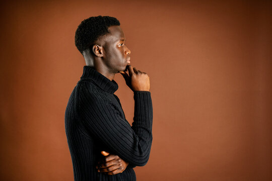Side view of pensive African American male rubbing chin and leaning on hand while standing on brown background in studio and looking away