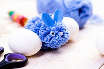 Fototapeta na wymiar This blue rabbit is made of pompons for Easter decor by hand. Easter bunny and white eggs on a stand Children's art project. DIY concept