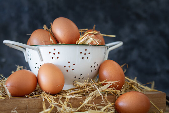Composition with brown chicken eggs in white bowl placed on wooden board with dry hay against dark background