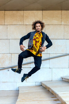 Full body of energetic young African American male in informal trendy outfit leaping on stairway against stone wall