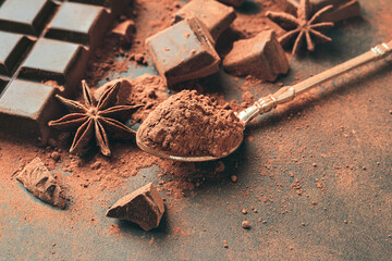 Brown cocoa powder in the spoon, chopped chocolate cubes and star anise on dark background