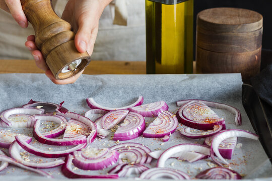 Unrecognizable chef with wooden pepper mill adding seasonings on chopped onions placed on baking paper on pan in domestic kitchen and cooking delicious dish