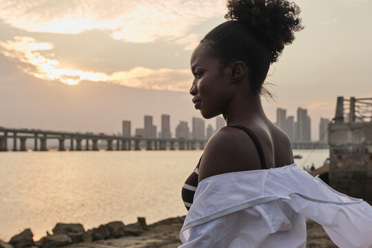 Tranquil pensive young African American female in casual clothes relaxing on urban seafront at sunset time