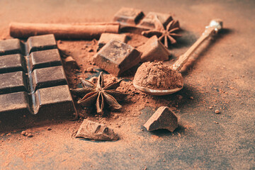 Brown cocoa powder in the spoon, chopped chocolate cubes, cinnamon stick and star anise on dark background