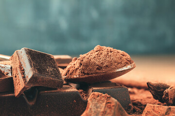 Brown cocoa powder in the spoon and chopped chocolate cubes, on dark background with copy space