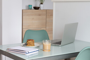 Empty table with laptop and notepad near prepare coffee and biscuits in light kitchen at home