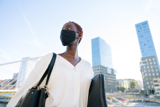 Low angle of stylish confident young African American businesswoman in black protective mask with handbag and laptop case standing on urban street with contemporary buildings