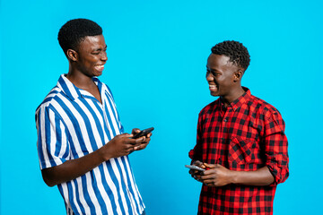 Content African American male best friends in trendy clothes browsing smartphones and laughing at jokes while standing on blue background in studio and looking at each other
