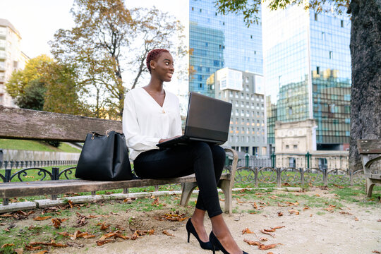 Positive young African American businesswoman in elegant clothes sitting on bench and working on laptop against blurred modern urban background