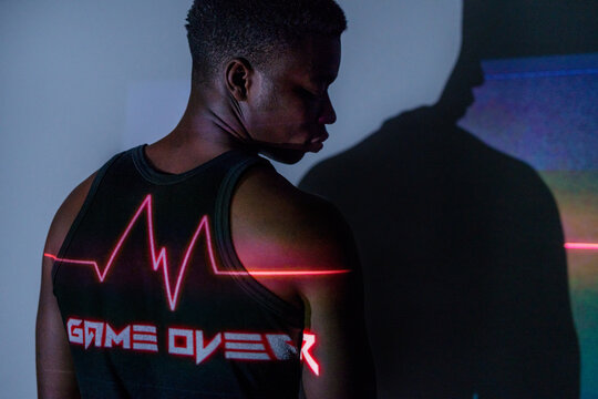 Back view of African American male standing in studio with red neon illumination of projector in shape of heartbeat line and inscription Game Over showing concept of end of life