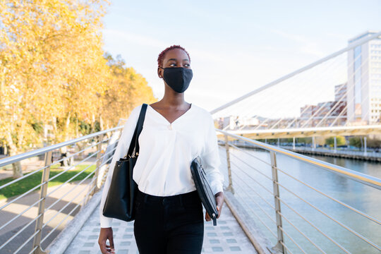 Front view of stylish confident young African American businesswoman in black protective mask with handbag and laptop case standing on urban street with contemporary buildings