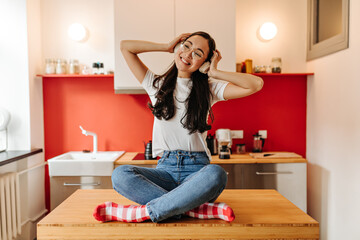 Lady in jeans and white T-shirt is sitting on wooden kitchen table and listening to music on headphones