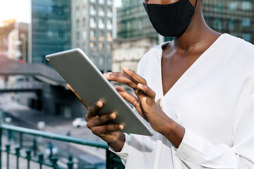 Crop of contemporary African American female entrepreneur in stylish outfit and protective mask browsing tablet while standing against modern glass building in city