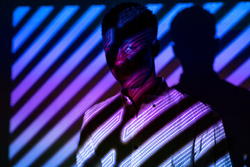 Unemotional African American male standing in neon light in shape of stripes in dark studio and looking at camera